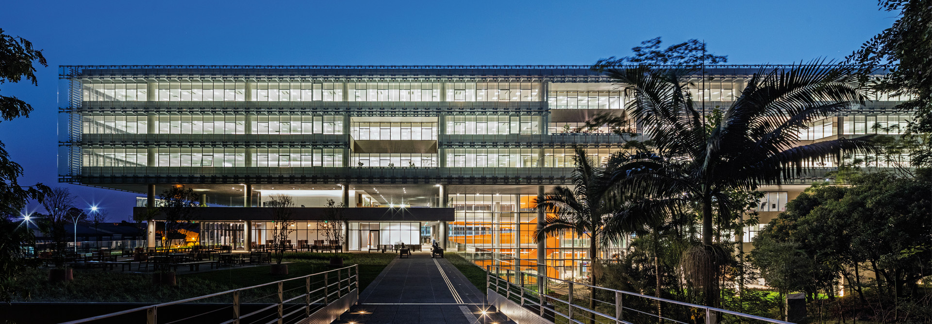 Fachada de um edifício corporativo moderno de vidro iluminado ao anoitecer, com passarelas de acesso e vegetação tropical ao redor.