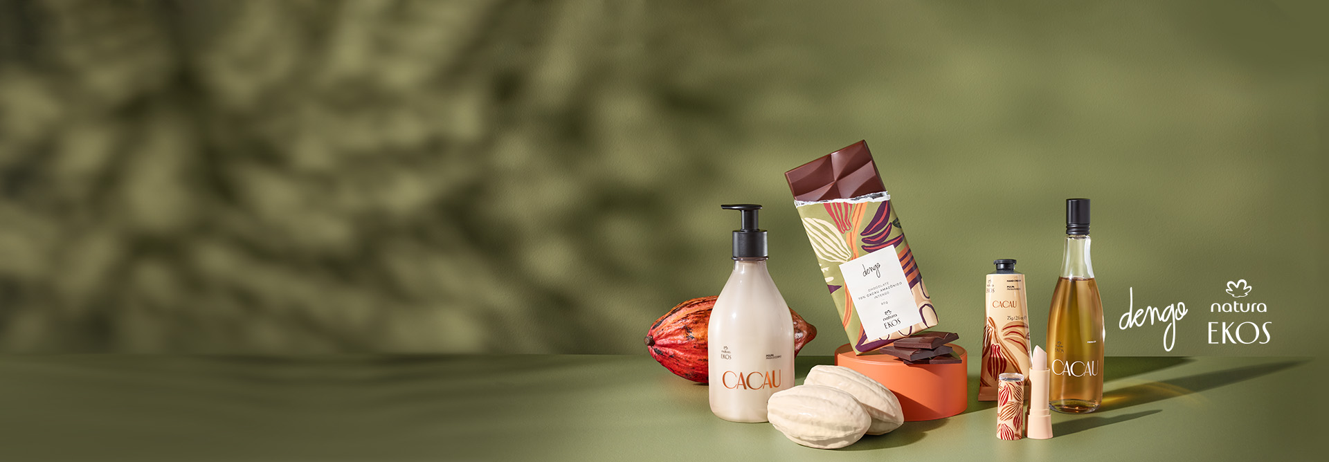 Foto traz fundo verde com sombras de árvore e produtos de Ekos Cacau, junto com barra de chocolate da dengo e frutos do cacau. Ao lado, está os logos de Natura Ekos e Dengo aplicados na imagem.