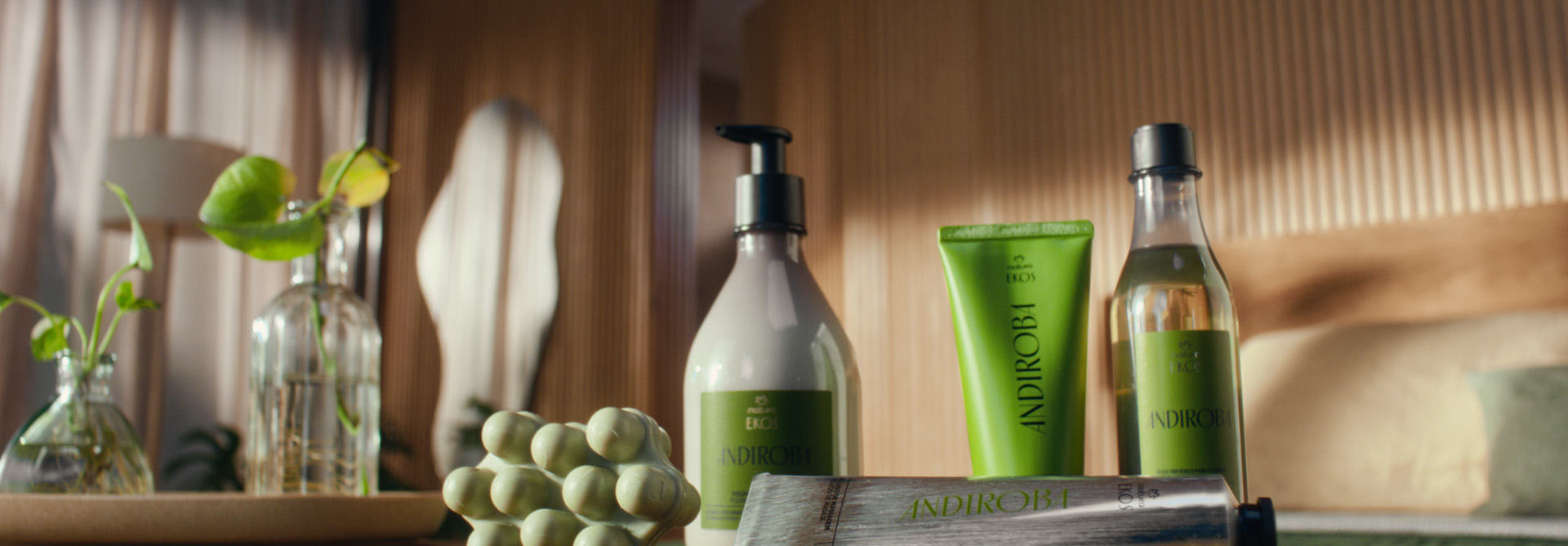 Vídeo de mulher em ambiente doméstico, utilizando os produtos da linha Ekos Andiroba. Finaliza com todos os produtos da linha e a flor Natura em textura fluída transparente em fundo verde-escuro.