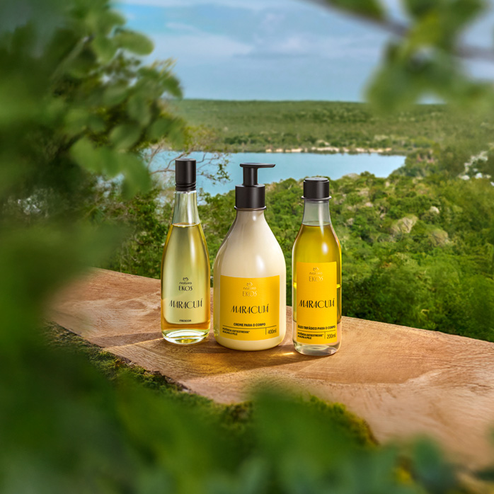 Três produtos Ekos Maracujá estão sobre uma superfície de madeira: óleo, creme para o corpo e sabonete líquido, todos com rótulo amarelo. Ao fundo, paisagem verde com lago.