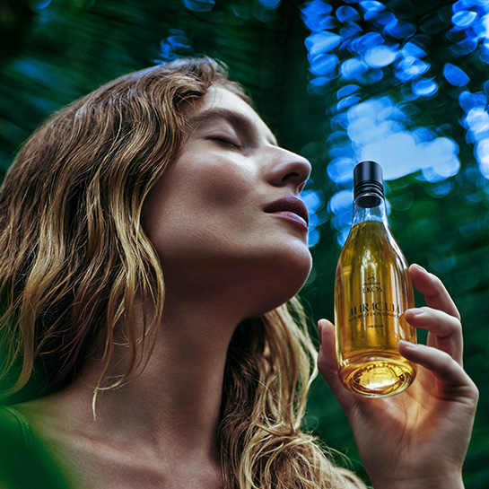 Mulher branca de cabelo loiro ondulado, de olhos fechados, segura um frasco com tampa preta e líquido amarelo do Ekos Frescor Maracujá próximo ao rosto. Fundo desfocado com vegetação densa.