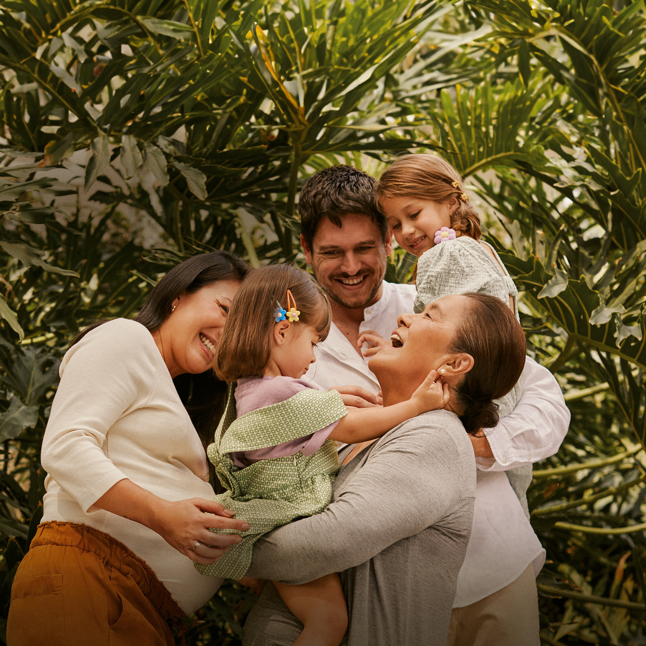 Família com 5 pessoas se diverte ao ar livre com plantas ao fundo. Uma mulher grávida, um homem, uma avó e duas meninas pequenas. 