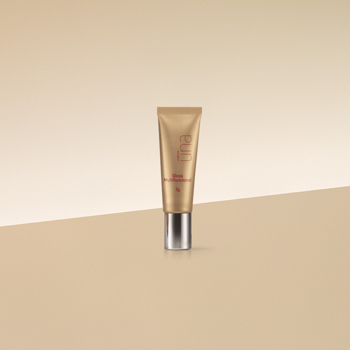 Bisnaga dourada com tampa prata de Gloss Multifuncional Natura Una Celebrar sobre bancada e fundo em amarelo-claro.