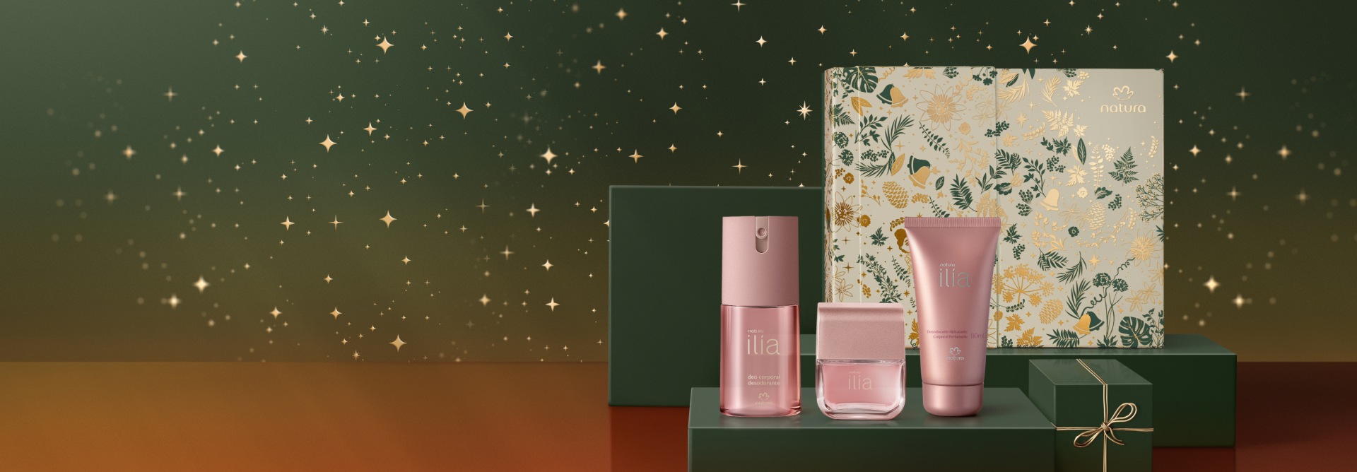 Presente Natura Ilía, com Deo Parfum, Desodorante Corporal e Desodorante Hidratante Corporal, em embalagens rosa perolado em fundo verde. Ao fundo, caixa decorada branca e dourada com estampa floral.