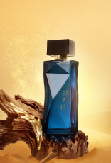Frasco de Essencial Oud Feminino, em tom de azul-escuro e tampa preta, aparece em destaque no centro da tela sobre fundo multifacetado dourado, fazendo alusão ao design do frasco de Essencial.