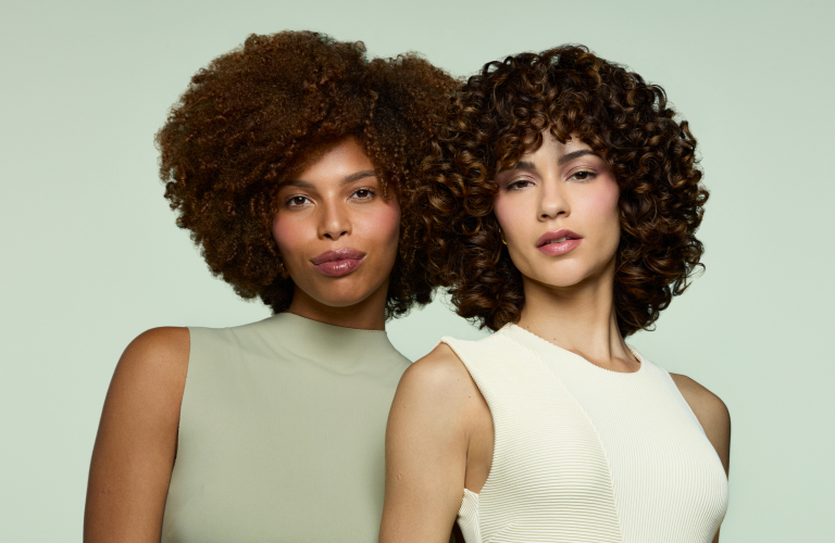 Foto de duas mulheres com cabelos crespos e cacheados castanhos posando juntas. Ao fundo, cor verde claro. Uma veste blusa verde e a outra, blusa branca.