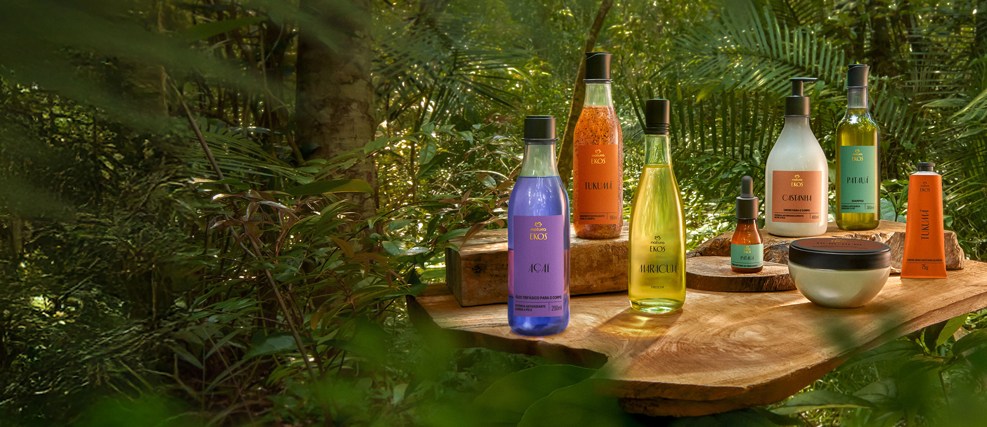 Uma variedade de produtos cosméticos da linha Ekos, dispostos sobre uma tábua de madeira rústica em um cenário de floresta ou jardim tropical. Os produtos incluem frascos e potes com líquidos e cremes de cores variadas (roxo, laranja, amarelo esverdeado, bege, verde claro) e rótulos com a marca 'Ekos', cercados por folhagens verdes e densas.