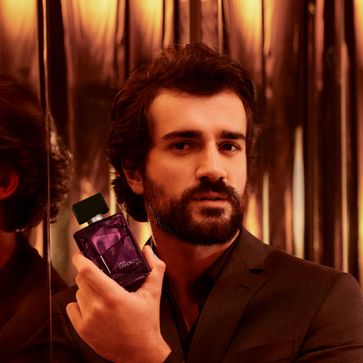 Homem branco com cabelos e barba castanhos e terno preto exibe Essencial Atrai Masculino em ambiente iluminado em tons de dourado e marrom.