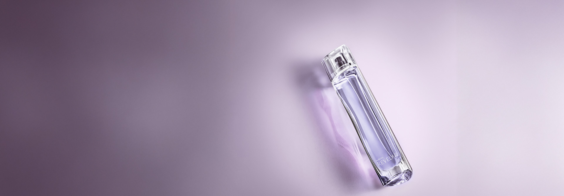 Desodorante Colônia Revelar Feminino, frasco transparente com líquido lilás. Ele está deitado diagonalmente sobre um fundo lilás suave, com uma sombra discreta.
