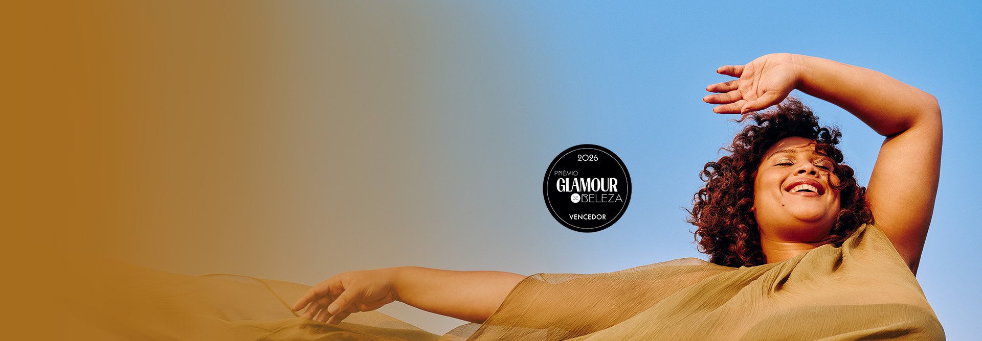 Uma mulher de pele parda e cabelos cacheados sorri com os olhos fechados sob a luz do sol, em um cenário ao ar livre com céu azul. Ela veste uma roupa leve em tom ocre que flutua ao vento. No centro da imagem, há um selo circular preto escrito "2026 Prêmio Glamour de Beleza Vencedor".