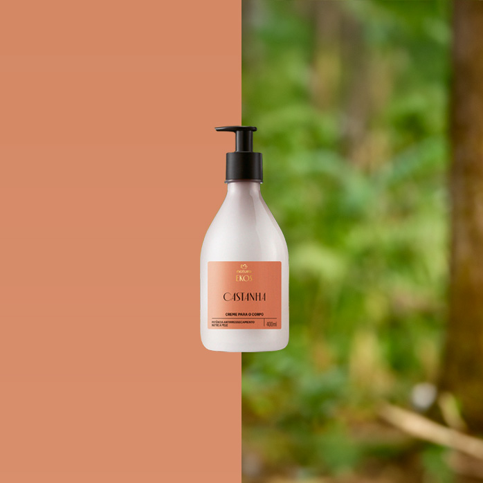 Frasco bege com rótulo marrom do Creme para Corpo Ekos Castanha, com válvula preta, está sobre tronco de árvore. Ao fundo, folhas verdes e troncos em ambiente de floresta.