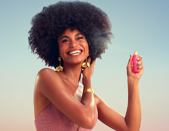 Modelo negra de cabelos crespos usa vestido longo rosê com brincos e bracelete dourados. Ela sorri para a câmera e borrifa o Perfume para Cabelos Luna Divina.