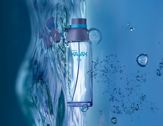 Frasco de Kaiak O2 feminino, em vidro translúcido, apoiado sobre água e respingos em fundo azul.