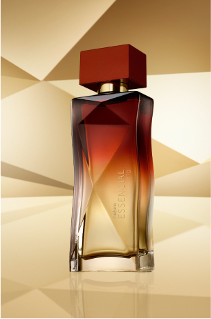 Frasco de Essencial Ato Feminino, nos tons de vermelho-queimado e dourado, aparece em destaque no centro da tela sobre fundo multifacetado dourado, fazendo alusão ao design do frasco de Essencial.