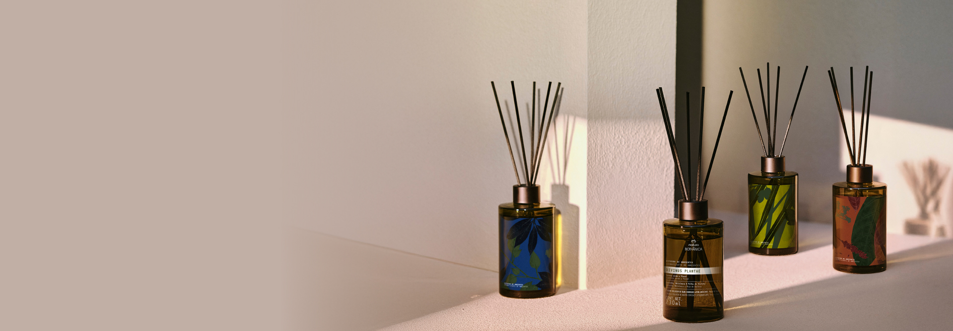 Novos Difusores de Ambientes Natura Bothânica aparecem em destaque em ambiente de paredes e superfície claras, com luz natural e leve sombra incidindo sobre os frascos.