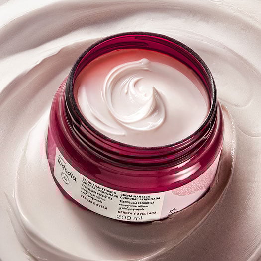 Creme Amanteigado Corporal Perfumado Tododia Cereja e Avelã em pote vermelho com creme branco. O pote está sobre um pouco da textura cremosa do produto espalhada ao fundo.