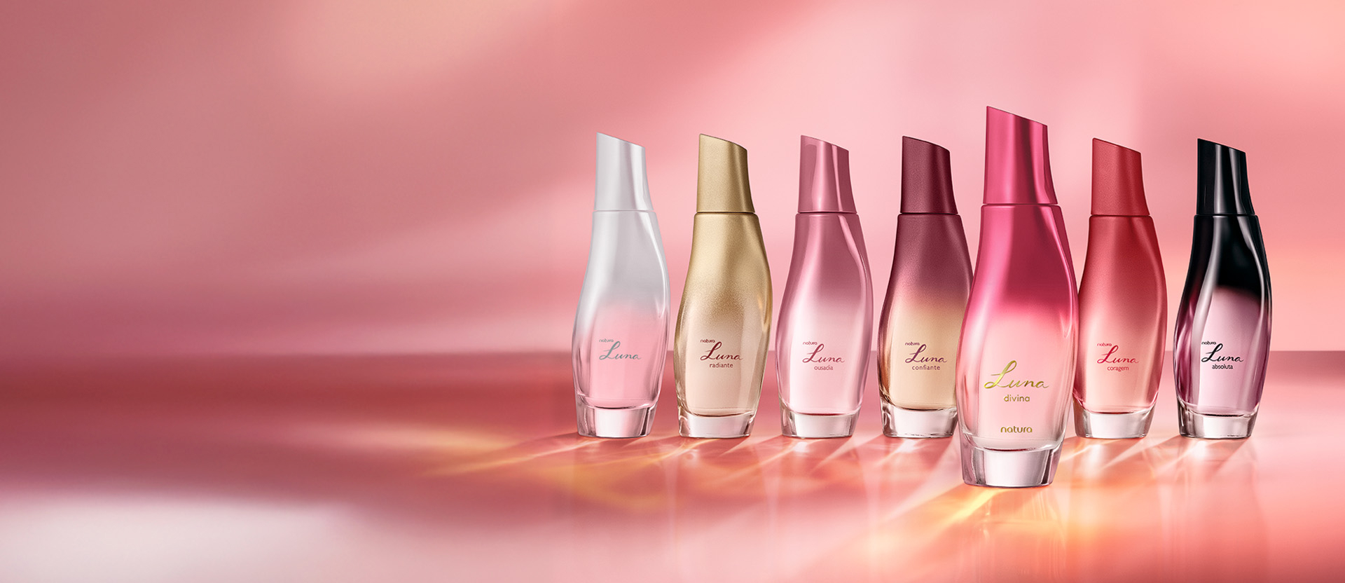 Sete frascos de Deo Parfums da linha Natura Luna alinhados. Os tons vão do branco, passando por dourado, rosa e vermelho, até o preto. O fundo é rosa degradê.