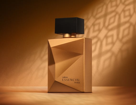 Deo Parfum Essencial Mirra Masculino em frasco facetado dourado com tampa preta. Está em fundo cor areia com sombra geométrica projetada.