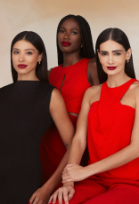 Três mulheres com diversos tons de pele usando batom vermelho intenso; duas vestem roupa vermelha e uma preta. Fundo neutro bege-claro, expressão serena e joias discretas.