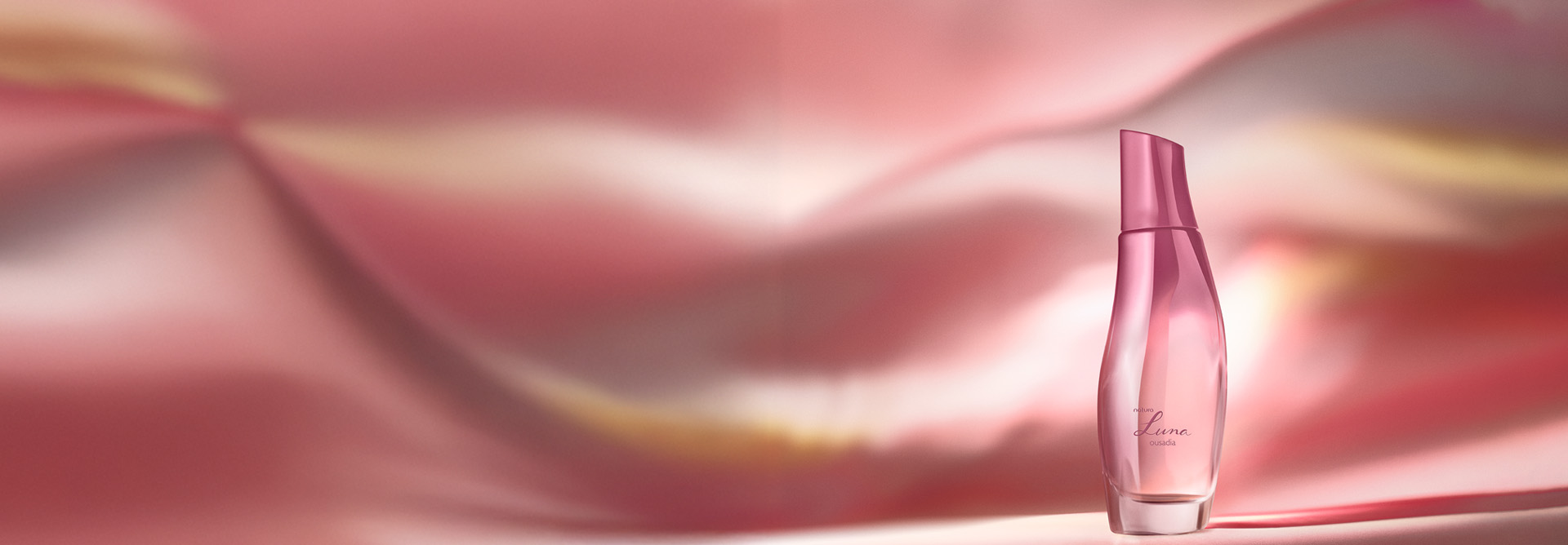 Desodorante Colônia Natura Luna Ousadia, com frasco rosado em acabamento fosco e transparente, destaca-se sobre fundo fluido em tons de rosa, pêssego e amarelo.