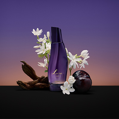Frasco roxo de fragrância Luna Nuit com flores brancas, madeira e frutas escuras. Fundo degradê roxo e rosado.