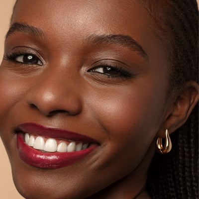 Close do rosto de uma mulher negra sorrindo, destacando uma pele iluminada e lábios com batom de tom vinho acetinado.