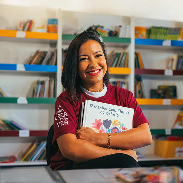 Professora com camiseta de Natura Crer Para Ver está abraçando um livro dentro da biblioteca de uma escola.