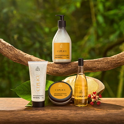 Itens Ekos Cupuaçu: bisnaga de Esfoliante, pote de Creme, frasco de Óleo Corporal e de Loção Corporal. Fundo com fruta de cupuaçu cortadas, com flores vermelhas e folhas verdes desfocadas.