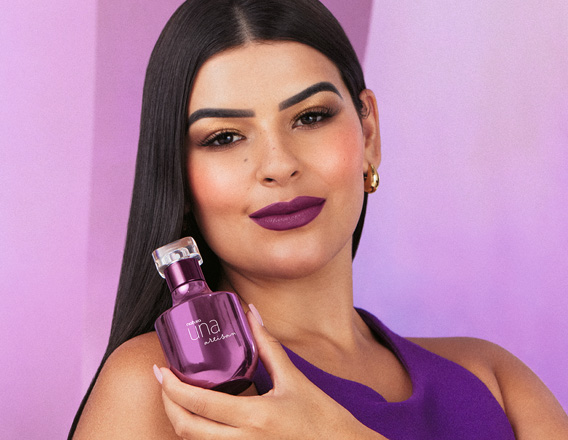 Foto de mulher de cabelos castanhos e longos, segurando uma fragrância Natura Una, em fundo lilás.