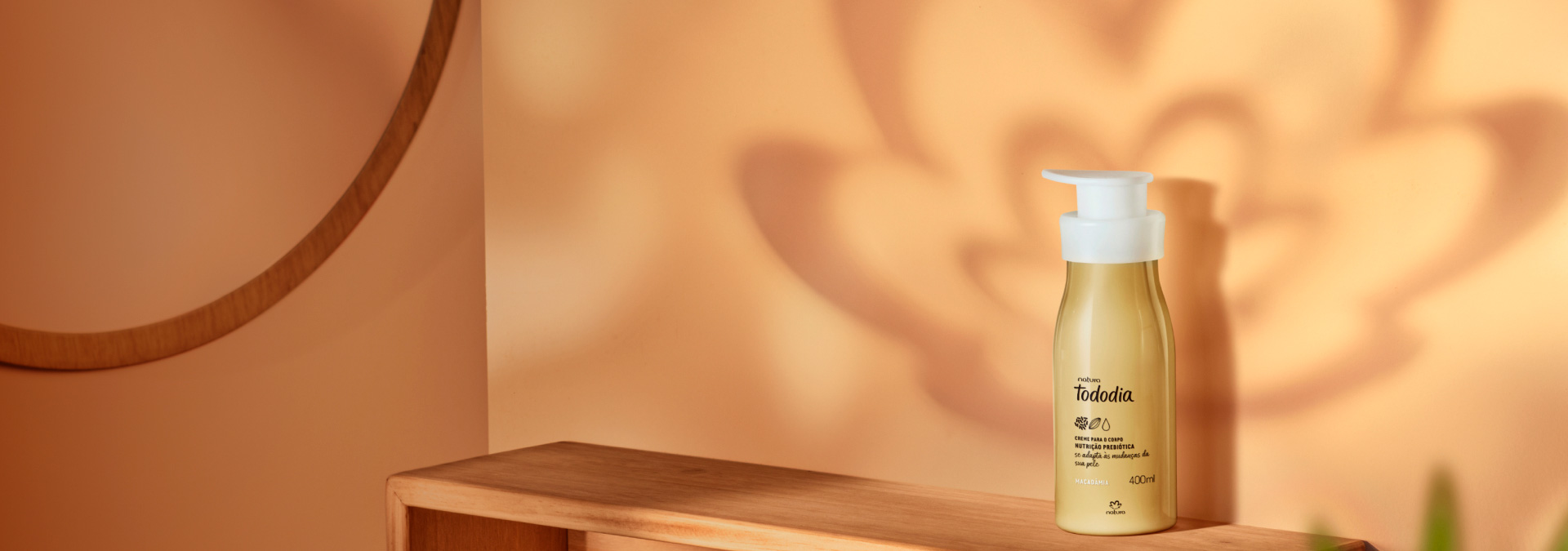 Hidratante para o corpo Tododia Macadâmia apoiado em cima de uma mesa refletindo a sombra da rosácea Natura.