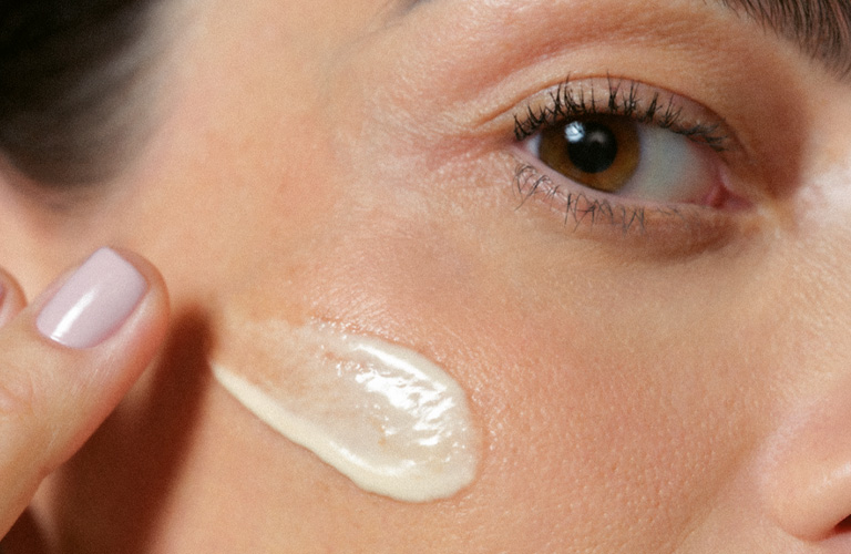 Foto com fundo neutro e rosto feminino em close-up, mostrando uma mão aplicando creme na bochecha com o dedo.