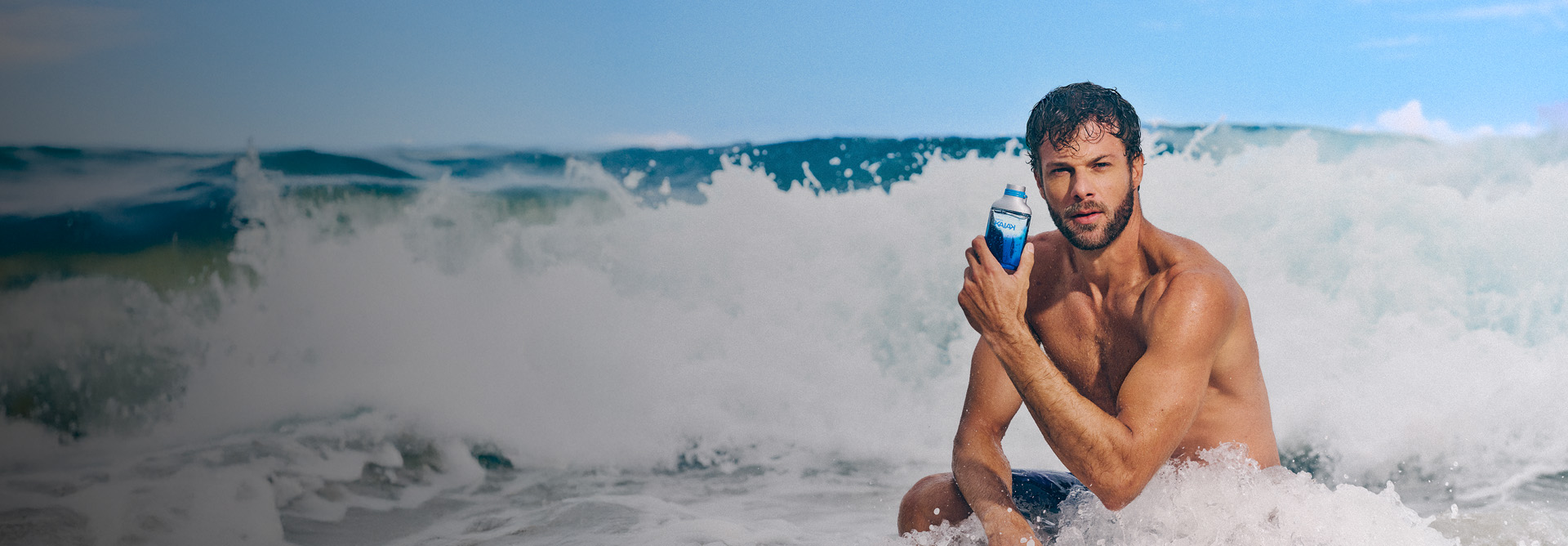 Homem sem camisa sentado na beira-mar com ondas quebrando. Ele segura frasco azul de fragrância Kaiak junto ao rosto.