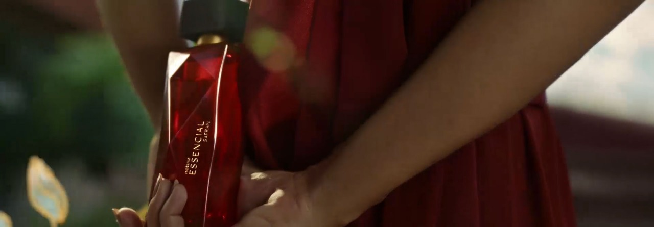 Mulher de pele morena e vestido vermelho em meio a um jardim. Ela segura delicadamente o frasco vermelho de Essencial Safran Feminino com uma das mãos atrás do corpo, como se guardasse um segredo. 