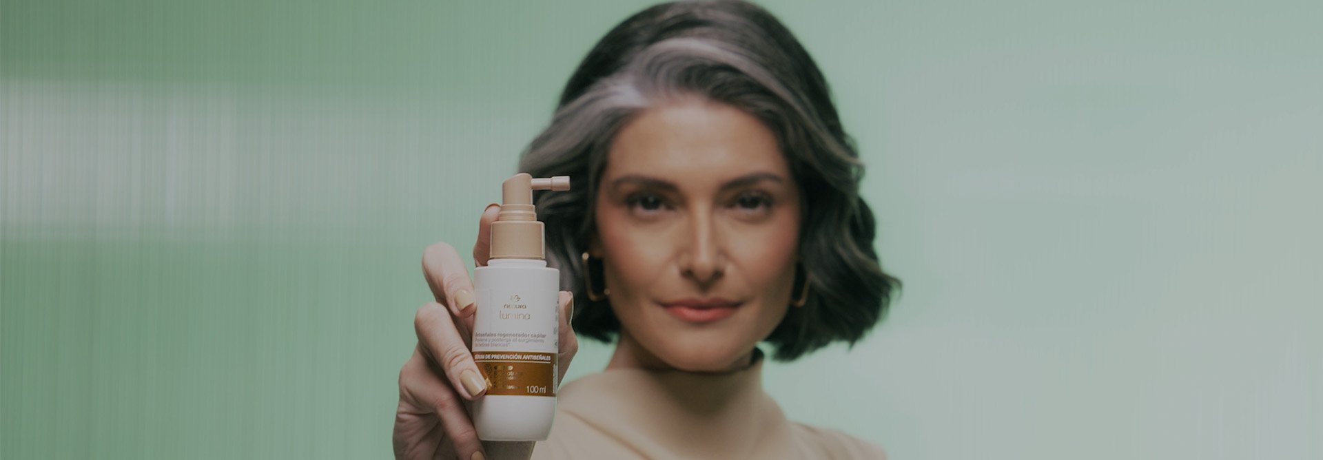 Vídeo com sequência de imagens: mulher branca de cabelos médios e grisalhos usando blusa bege, imagem de produtos da linha Natura Lumina Antissinais, mulher aplicando itens nos cabelos, animação de gota dourada e sua ação no couro cabeludo e fios de cabelo. Vídeo finaliza com mulher segurando item próximo ao corpo, em fundo verde.