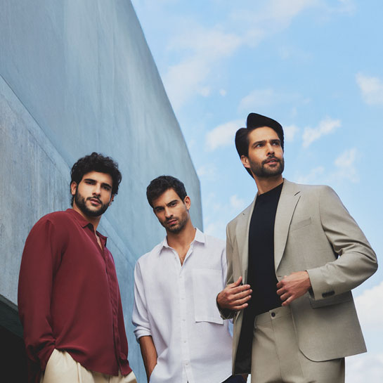 Três homens jovens de cabelos e barbas castanhos, à esquerda de camisa vinho, de camisa branca no centro e à direita de terno cáqui e camisa preta, em fundo de concreto e céu azul.