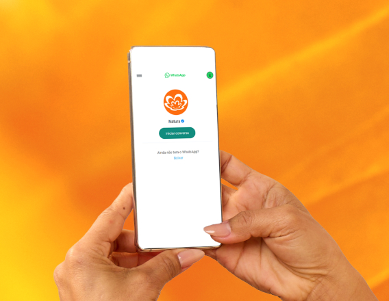 Consultora de Beleza Natura e Avon, de pele morena e cabelos escuros e curtos, está sentada à mesa escrevendo. À sua frente, revistas Natura e Avon, app Natura no celular e site Natura no notebook.