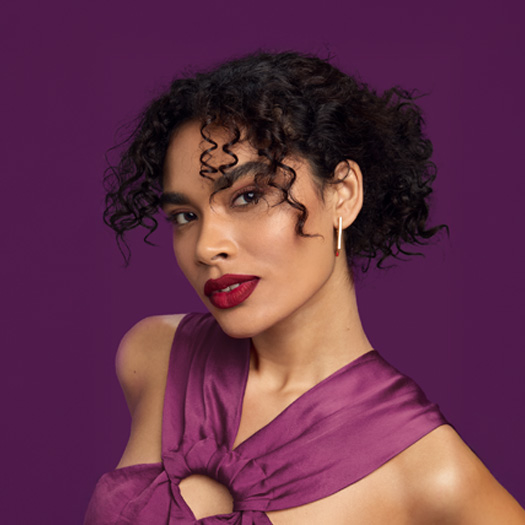 Mulher de cabelos ondulados e presos, maquiada e usando blusa de alças roxas em fundo roxo.