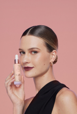 Foto de mulher com cabelos presos e loiros segurando um produto Natura Una próximo ao rosto, em fundo rosa-claro.