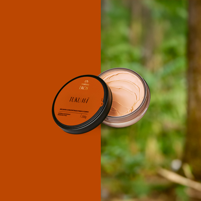 Pote aberto do Bálsamo Concentrado para o Corpo Ekos Tukumã, tampa laranja e creme bege denso. Fundo dividido entre laranja sólido e floresta desfocada ao fundo.