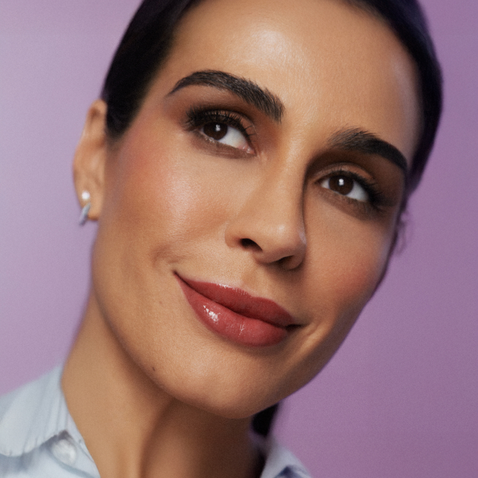 Modelo morena de cabelos presos sorri com o Gloss Labial Natura Una nos lábios enquanto olha para o canto esquerdo da tela.