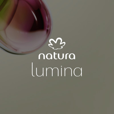 Logo de Natura Lumina, em letras brancas, destaca-se no centro da tela sobre textura aquosa em formato de gota nos tons de roxo, amarelo e verde. Fundo acinzentado.
