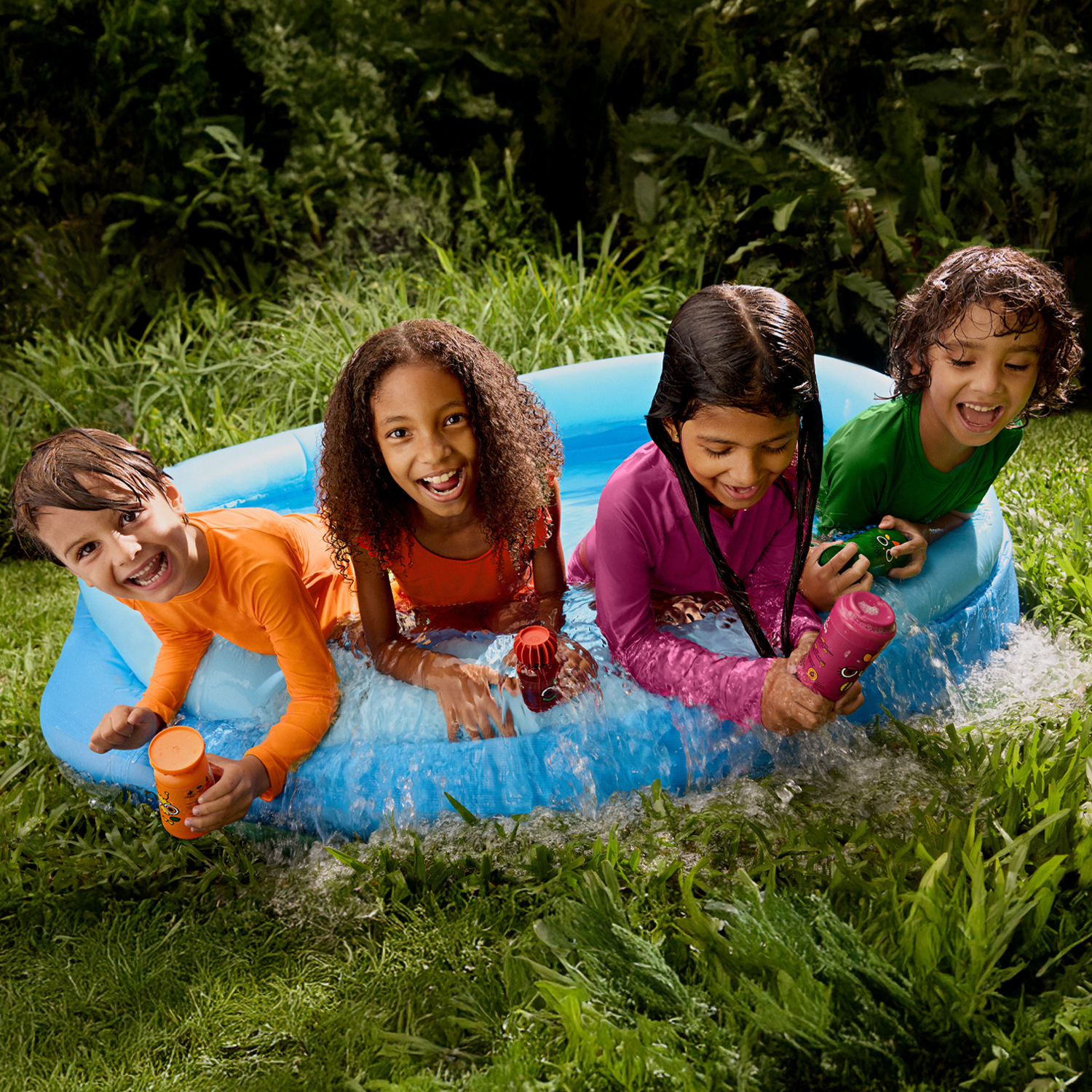 4 crianças vestindo roupas coloridas estão dentro de uma piscina inflável, são 2 meninos e duas meninas. A piscina está em um campo verdinho e as crianças seguram produtos de Naturé. 