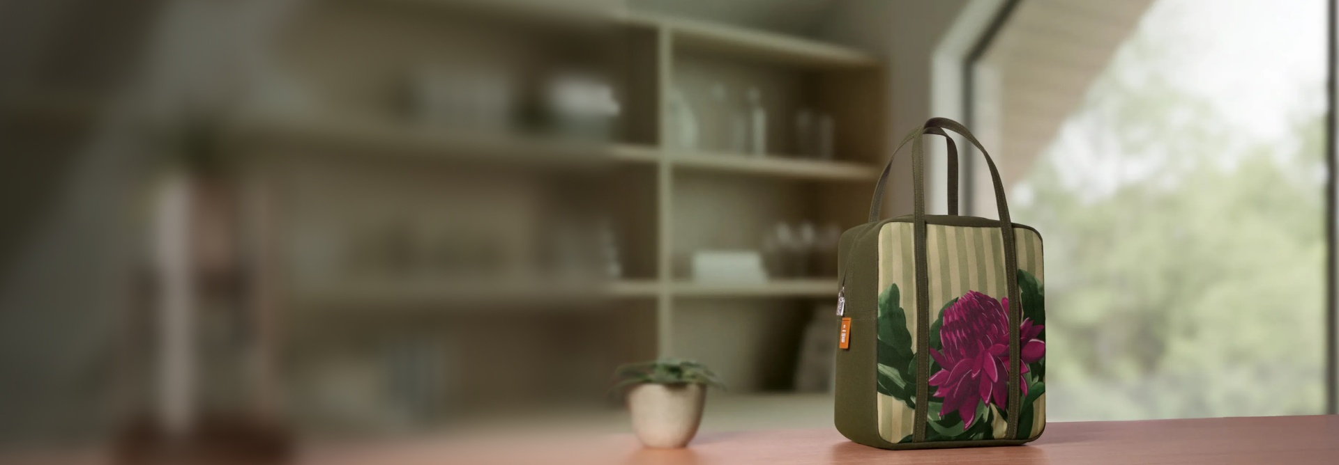 Bolsa Crer Para Ver em tecido verde-oliva, estampada com uma flor roxa de folhas verdes. Possui alça de mão verde e está sobre um armário de madeira. Fundo desfocado que lembra ambiente de cozinha.