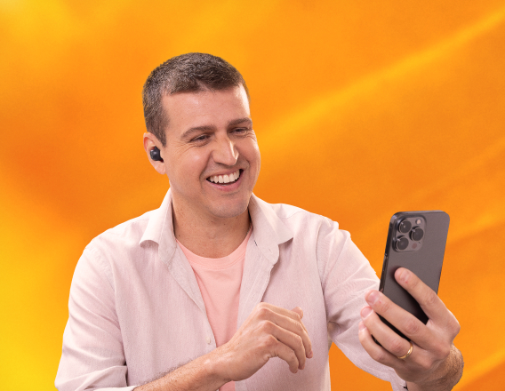 Imagem mostra homem de pele clara e cabelos curtos e castanhos vestindo camiseta rosada sobreposta por uma camisa branca. Ele usa fones de ouvido sem fio e sorri diante do celular. Fundo amarelo.