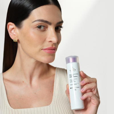 Mulher branca segura produto Chronos Derma próximo ao rosto. Ela usa regata rosada, cabelos presos e está em fundo claro.