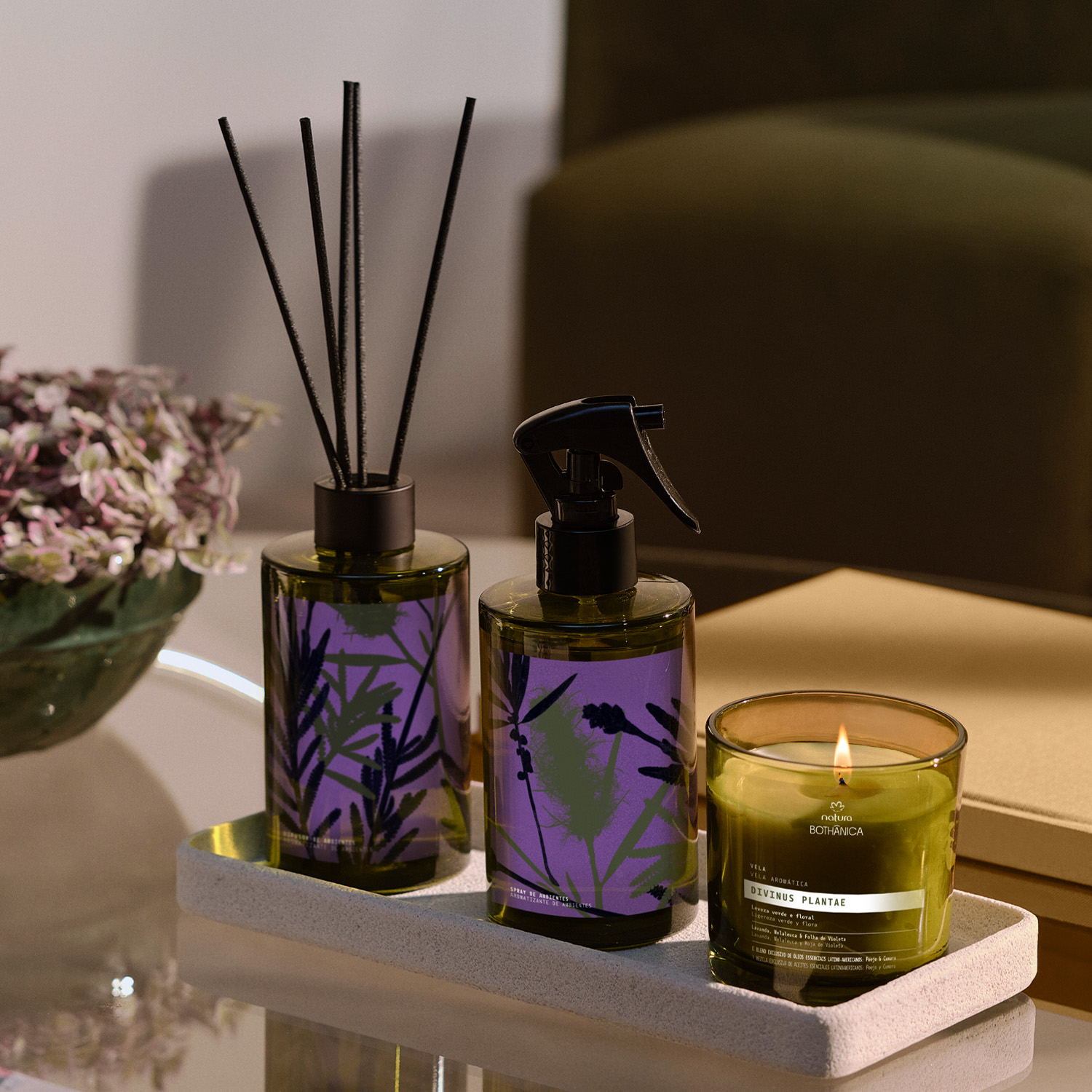 Bandeja de pedra sobre uma mesa de centro branca com produtos Bothânica Divinus Plantae: 1 difusor de ambientes, 1 spray e uma vela aromática. Embalagens de vidro verde com ilustrações em roxo.