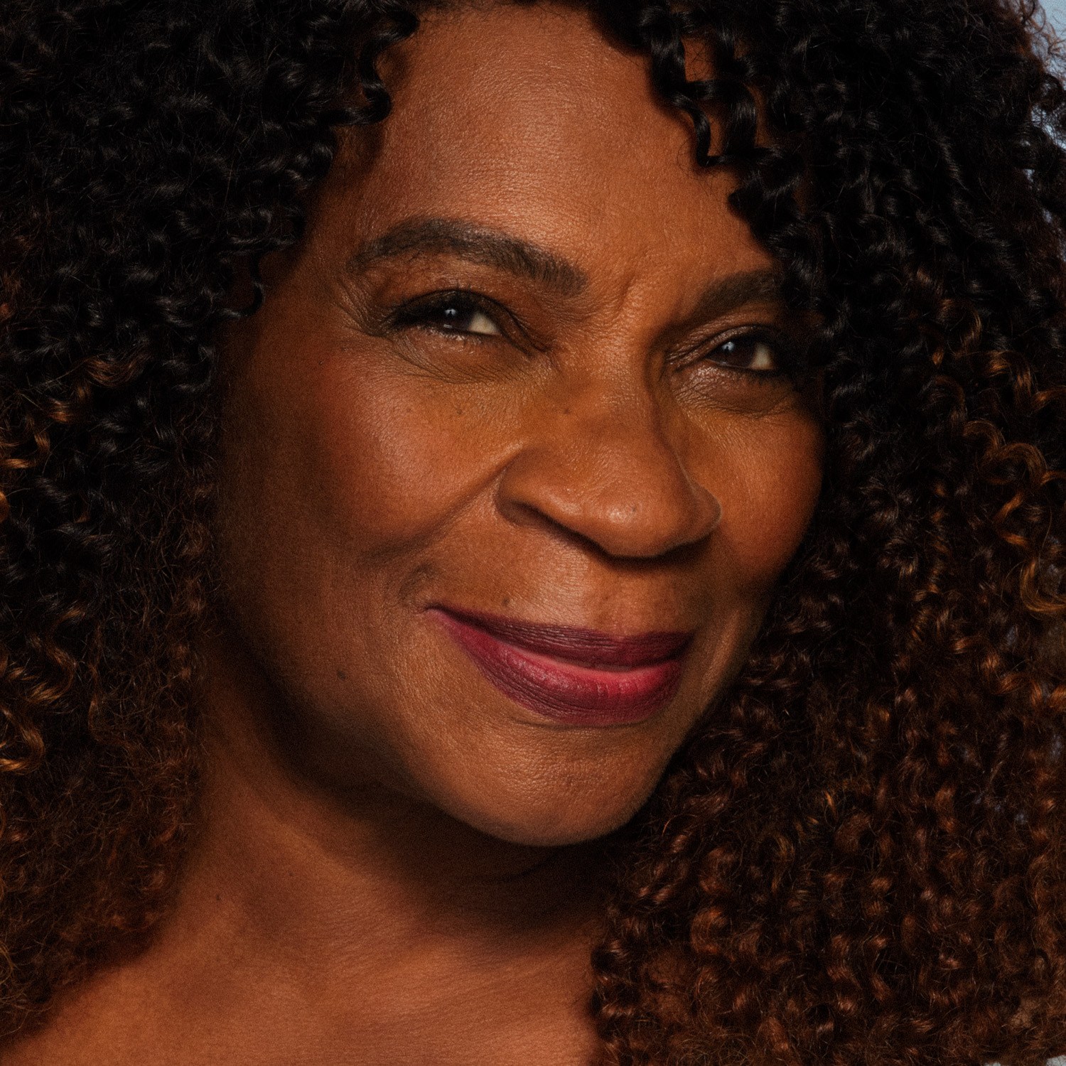 Foto de uma modelo de pele negra e cabelos crespos sorrindo. Ela está usando maquiagem leve, com batom roxo.