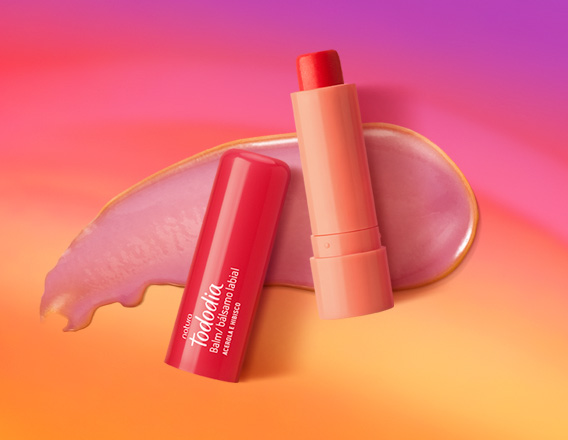 Embalagem rosada aberta de Balm Labial Tododia Acerola e Hibisco sobre textura rosada cremosa, em fundo em tons de laranja e rosa.