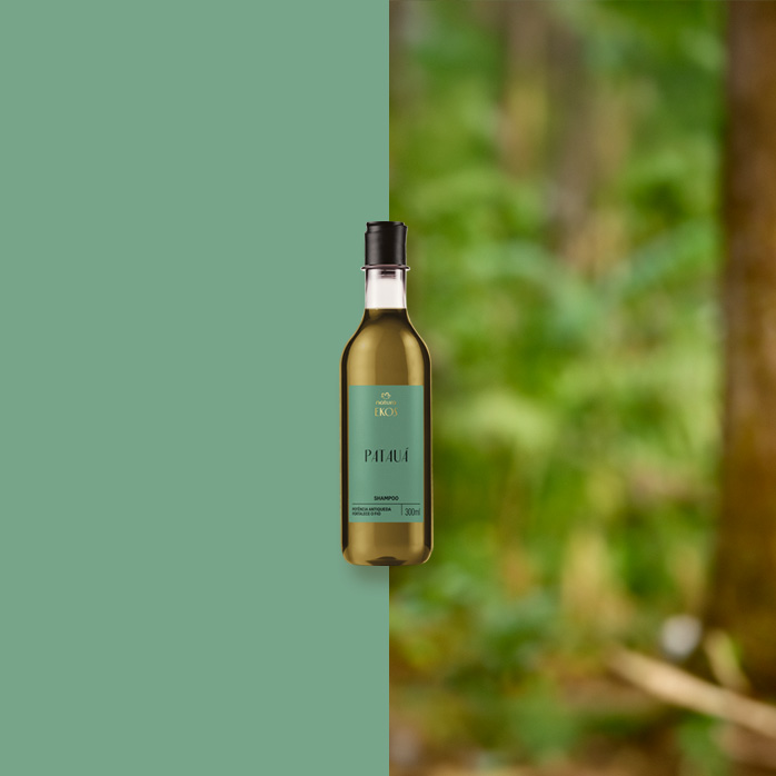 Frasco transparente de Shampoo Ekos Patauá com rótulo verde e tampa preta está centralizado entre fundo verde e floresta desfocada, transmitindo conexão com a natureza.
