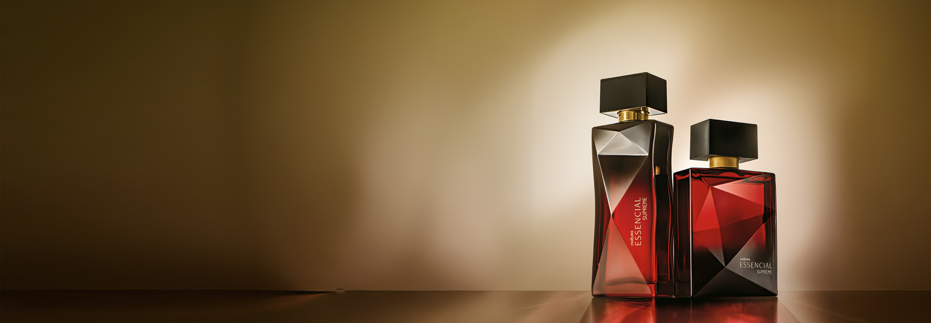 Essencial Supreme Feminino e Masculino, com frascos em tons de vermelho e preto e tampas pretas, aparecem iluminados no canto direito da tela sobre superfície e fundo em tom de marrom.